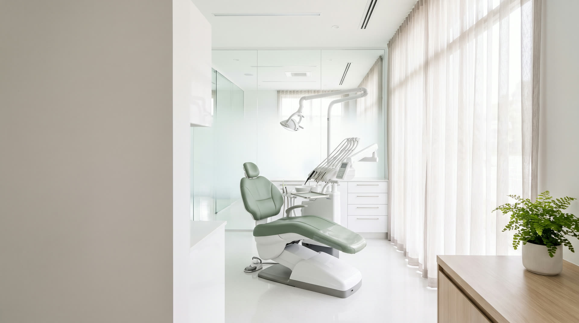 A bright dental treatment room at Cedar Grove Dental with a sage-green chair, sheer curtains and a small fern